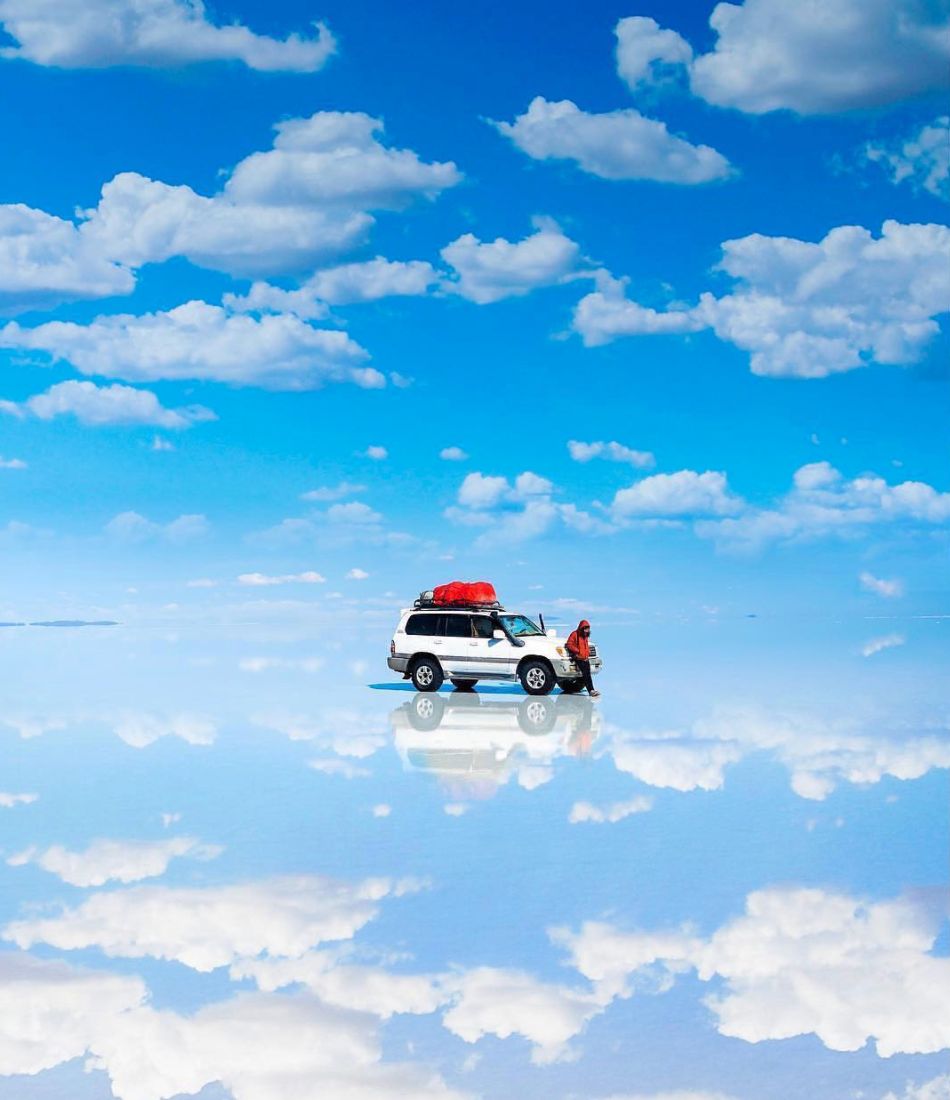 A tour jeep and a traveler reflected in the flooded Salar de Uyuni salt flats under a sky full of clouds, Bolivia