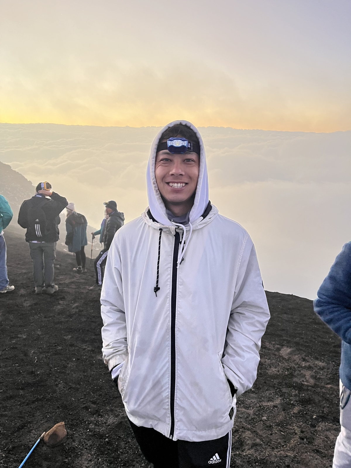 Jonathan Byrne wearing a white hooded jacket and headlamp, smiling above a sea of clouds at sunrise